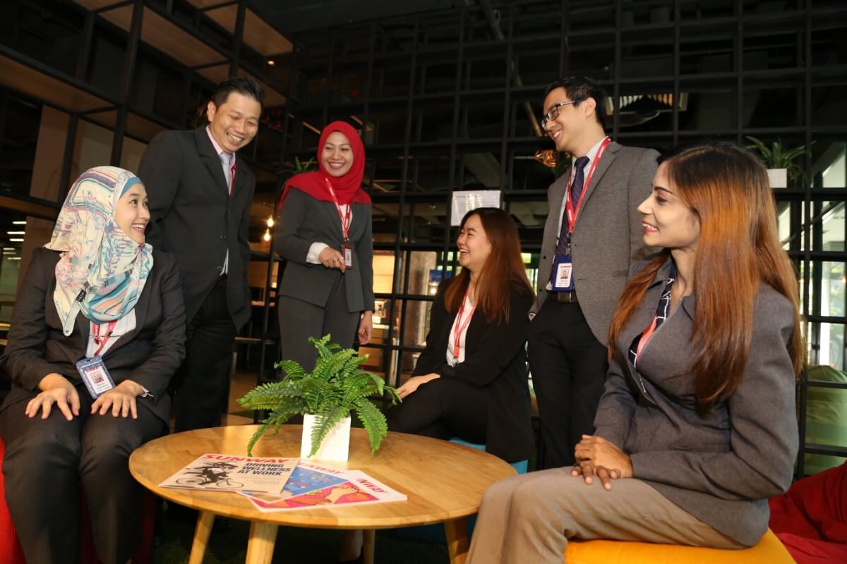 A medium wide shot of Sunway’s multiracial employees interacting cozily round a coffee table, against a dark backdrop