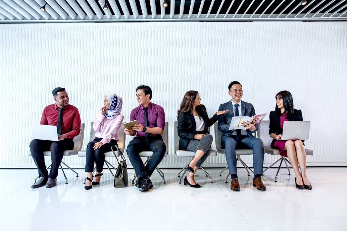 A wide shot of Sunway’s multiracial employees sitting side-by-side, interacting with one another, against a bright backdrop