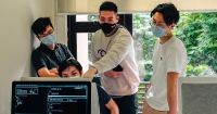 A mid-shot of four male students huddled in proximity while wearing face masks, facing a computer screen at 42KL against a large window displaying the lush bamboo garden outside