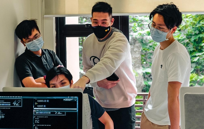 A mid-shot of four male students huddled in proximity while wearing face masks, facing a computer screen at 42KL against a large window displaying the lush bamboo garden outside