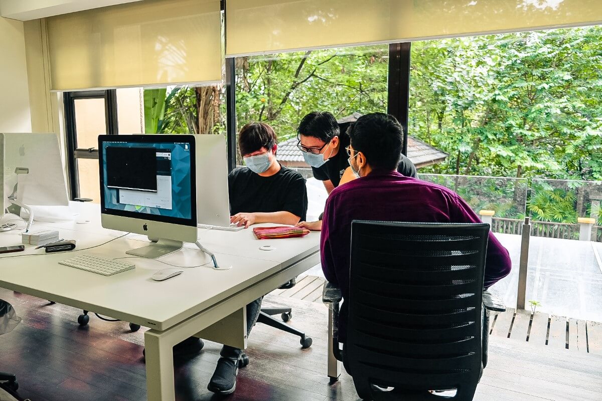 A mid-shot of three male students at 42KL, huddled closely facing a computer screen discussing around a computer, all of them wearing face masks