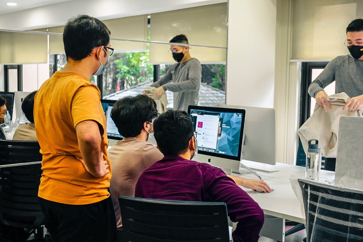 A mid-shot of four male students at 42KL, with three of them huddled closely facing a computer screen with their backs facing the camera while the other stood opposite them, all of them wearing face masks