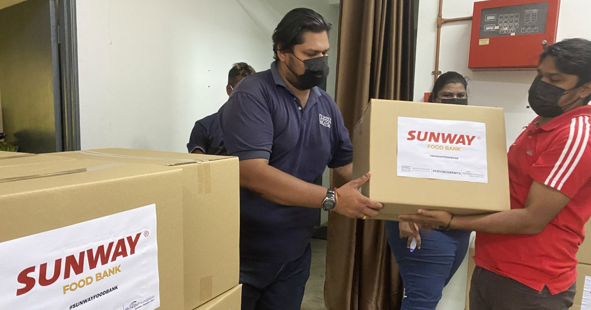 A medium wide shot of a burly male in navy outfit passing a carton box with a Sunway Food Bank label printd upfront to an equally burly male in red top and charcoal grey pants while a lady in denim witnesses the exchange