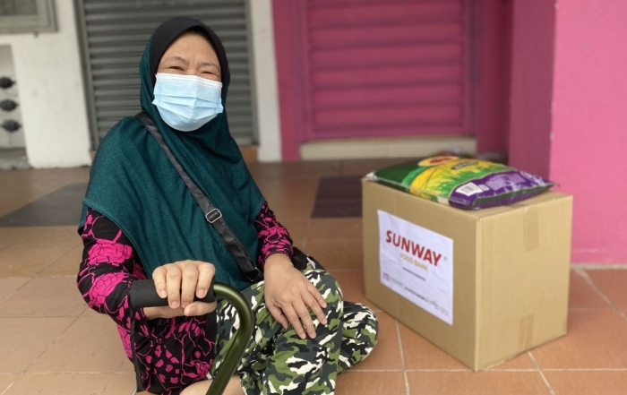 A wide shot of a happy female with a walking stick, sitting on steps leading towards some shop corridors with a #SunwayforGood food bank and a bag of rice resting atop it by his side