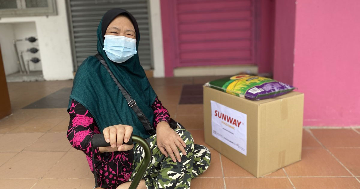 A wide shot of a happy female with a walking stick, sitting on steps leading towards some shop corridors with a #SunwayforGood food bank and a bag of rice resting atop it by his side