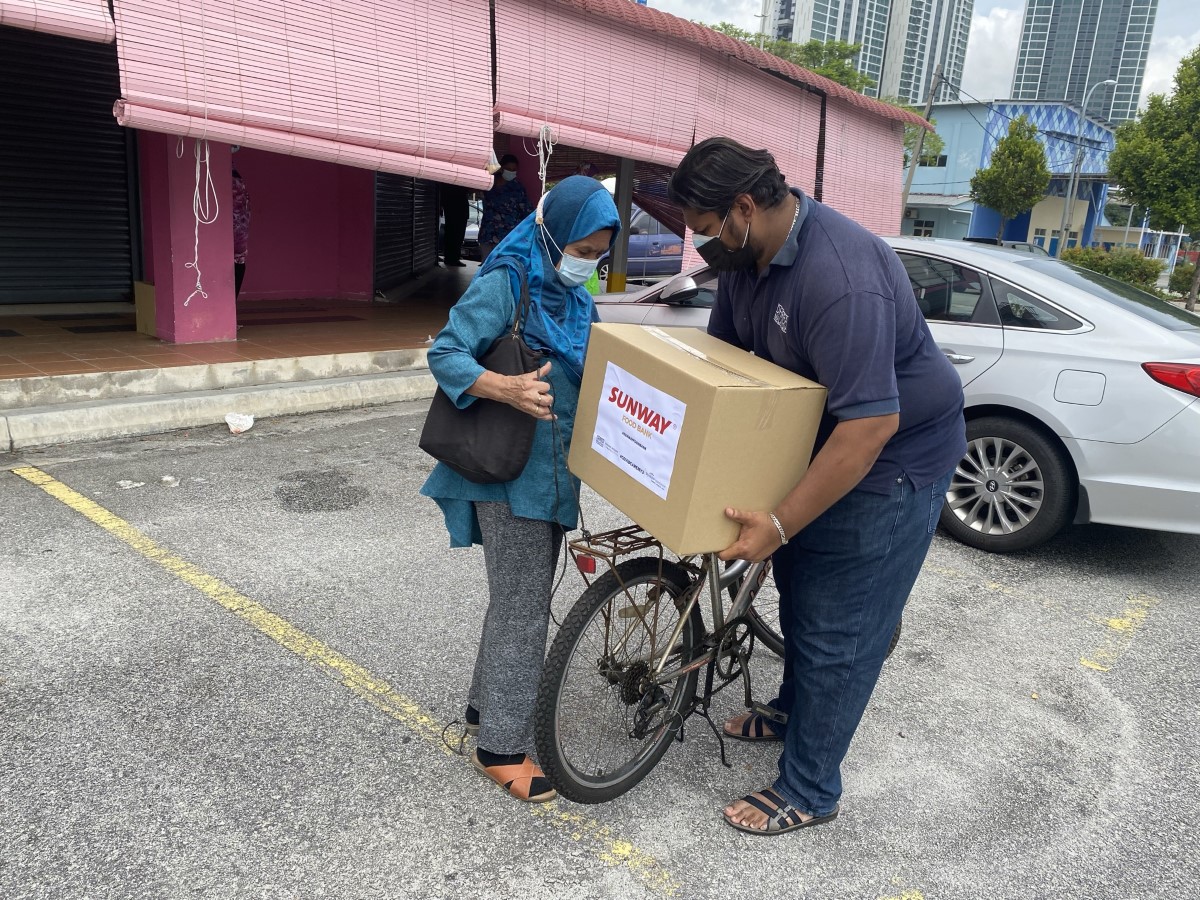 A wide shot of a dark-skinned burly male propping a #SunwayforGood food bank box on the back of a bicycle as a lady dressed in bright cobalt blue headscarf and top looks on