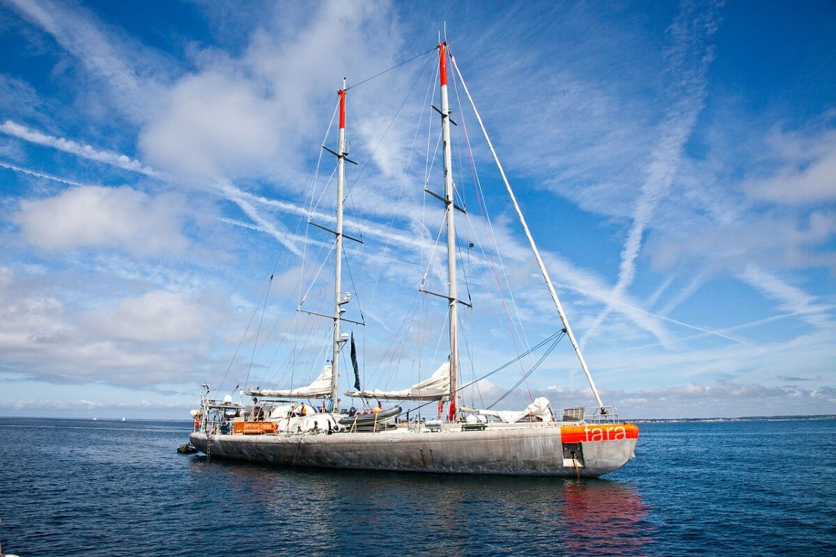 A wide-shot of a white schooner with minimal orange accents in the middle of the ocean during daytime, with multiple contrails on an otherwise clear sky