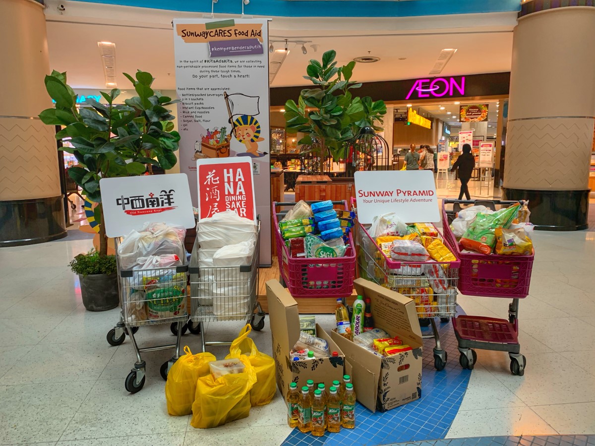A wide shot of trolleys and boxes of processed dry food gathered neatly together with a Sunway Pyramid Mall sign propped up amidst the goods and a SunwayCARES Food Aid banner hoisted in the background