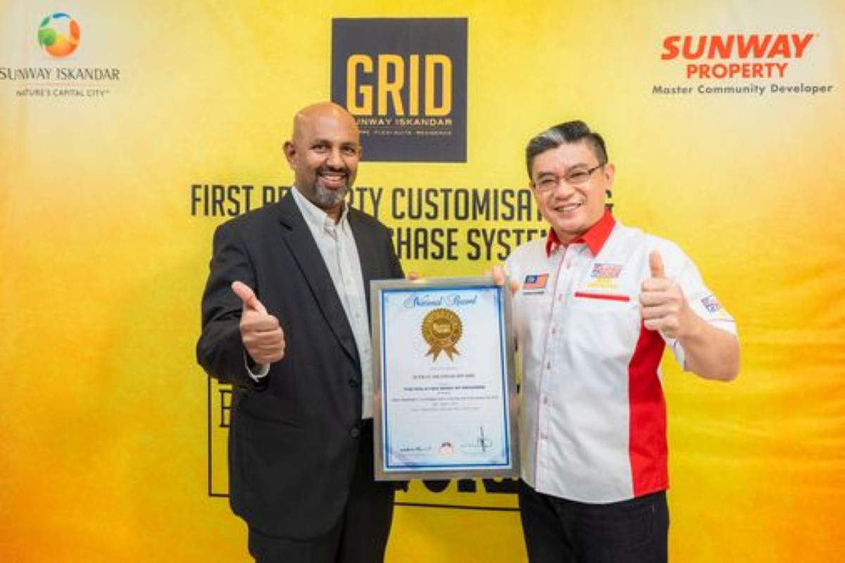 A mid shot of two males, each stood holding one side of a framed certificate with their thumbs up, looking visibly pleased in front of a yellow backdrop featuring Sunway GRID, Sunway City Iskandar Puteri and Sunway Property logos.