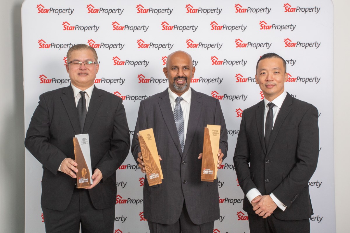 A mid shot of three males dressed smartly in matching suits and expressions, with the first two gentlemen holding up uniquely-shaped trophies as the third male stood by with his hands clasped, with a white backdrop for The StarProperty Awards behind them.