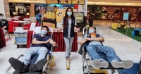 A wide shot of two males in surgical masks propped up on recliner beds, placing light pressure on the crevices of their elbows, while a female in surgical mask flashes two thumbs up in between them