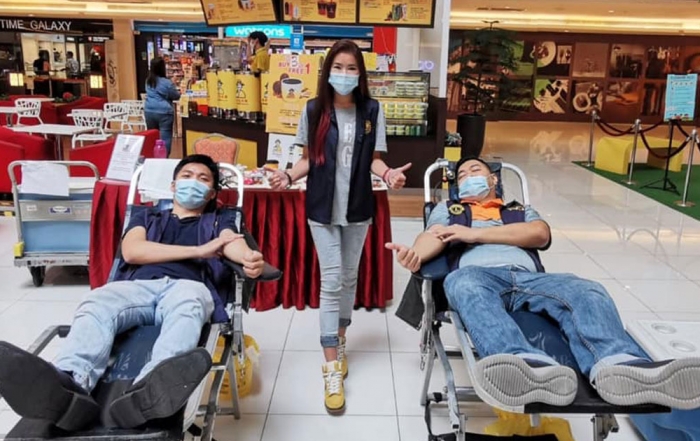 A wide shot of two males in surgical masks propped up on recliner beds, placing light pressure on the crevices of their elbows, while a female in surgical mask flashes two thumbs up in between them