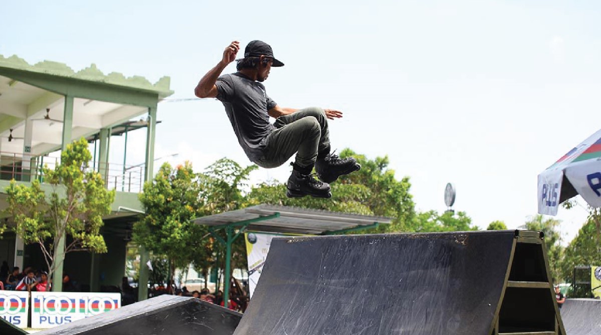 A wide shot of a male in action midair, dressed in charcoal grey ensemble, wearing a snapback and a pair of black rollerblades, cruising off a ramp.