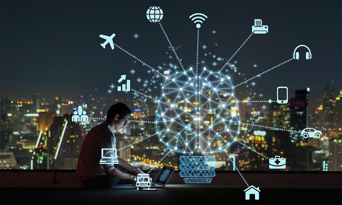 A wide shot of a sitting male talent under an open night sky, face illuminated by his open laptop as a huge hologram is projected over his person and vicinity, with a lit city skyline in the background