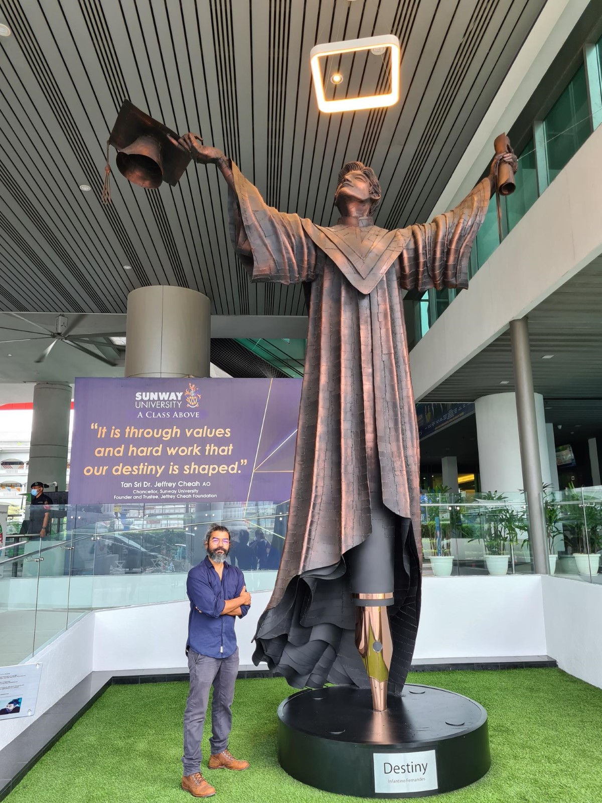 A portrait shot of Sunway’s resident artist, Infantino Fernandes, standing tall with both arms crossed next to the sculpture ‘Destiny’