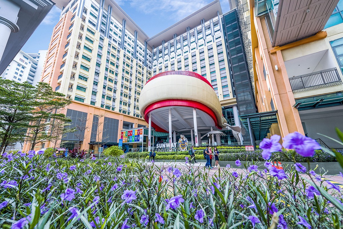 A low-angle full landscape shot of Sunway University’s vibrant and open campus during daytime, featuring impeccably-designed infrastructures and nature elements, with a prominent signboard displaying United Nation’s 17 Sustainable Development Goals amidst the sophisticated ambience