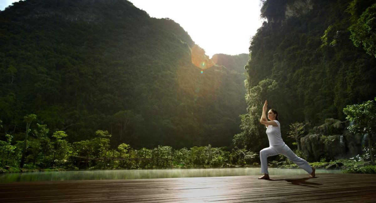 A wide angle shot of a woman in white practicing yoga in the pristine, open space within The Banjaran Hotsprings Retreat in Ipoh, Perak, bathed by warm, gentle sunrays.