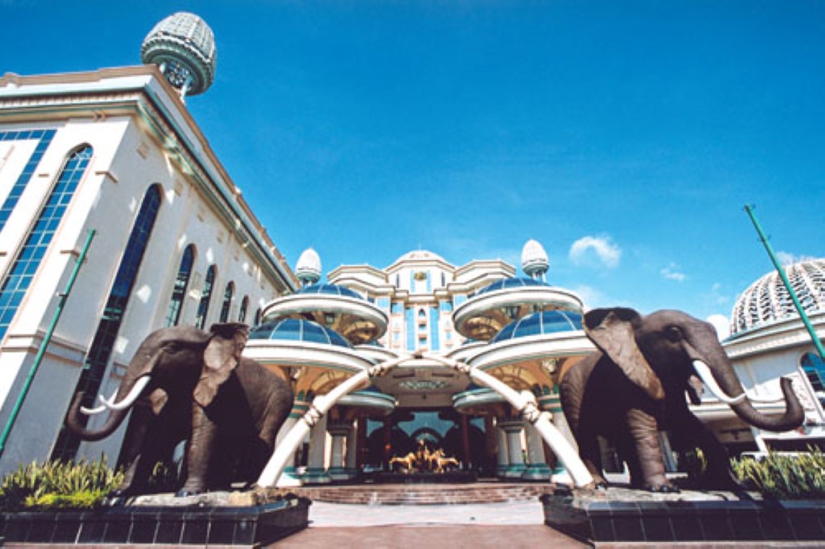 A low angle entrance shot of Sunway Resort circa 1996, flanked by an elephant on each side, its white façade with golden trims and bright blue glasses appears grand and majestic under the bright, blue sky.