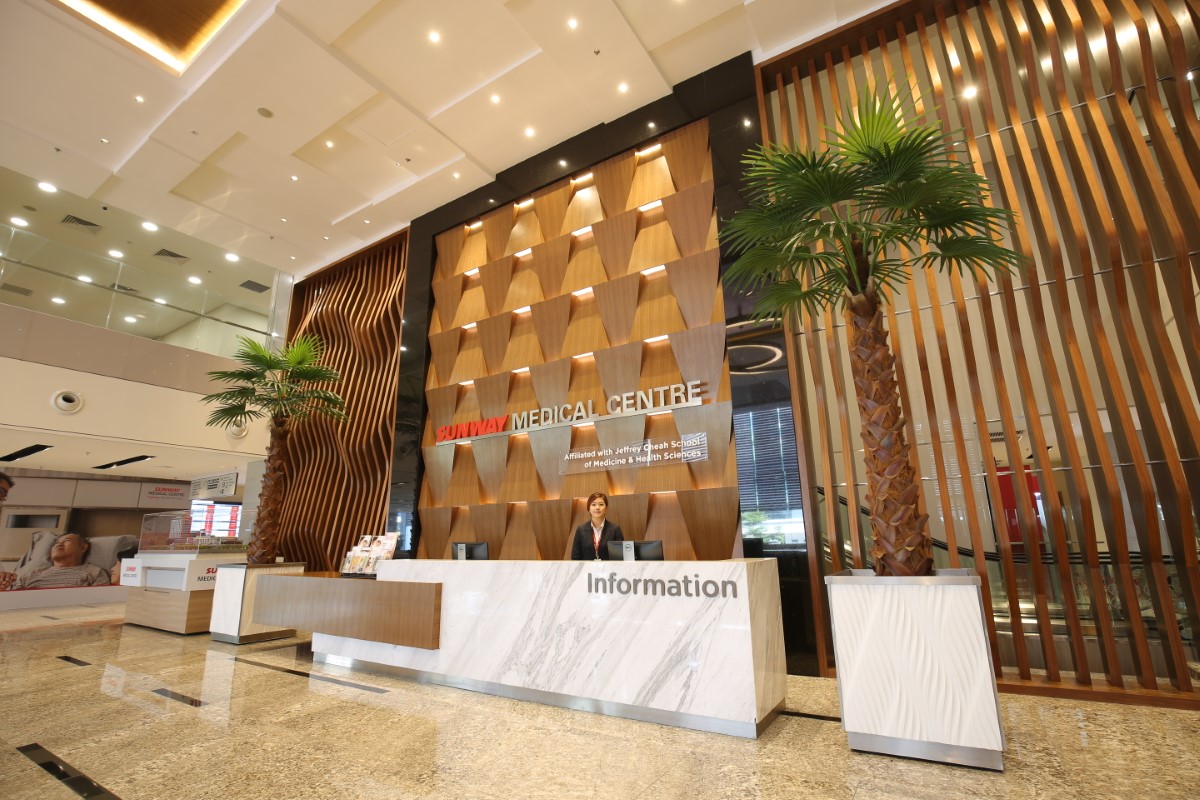 A close-up view of present-day Sunway Medical Centre, its statement wall now an accented wooden wall with a refreshing, modern twist, the marbled reception counter now surrounded by two pots of tall palm trees to bring the outdoors, indoors.