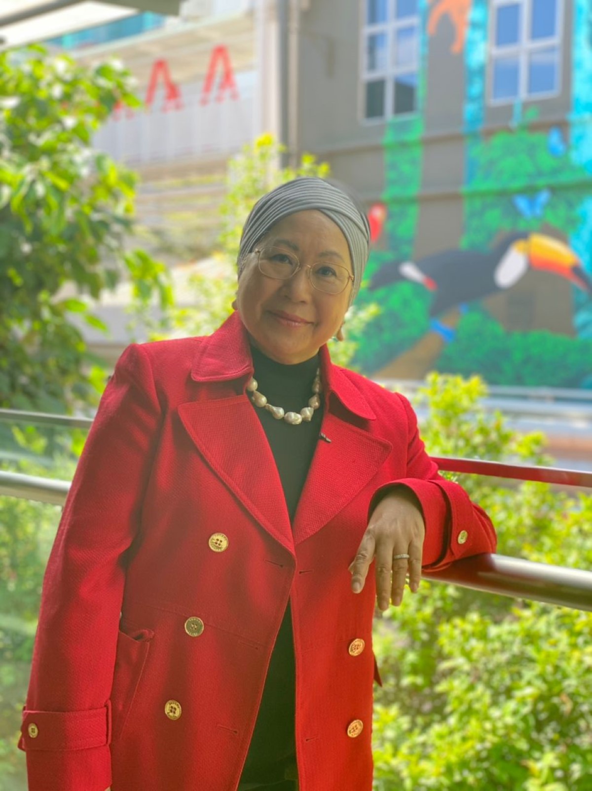 A mid-wide shot of Tan Sri Dr. Jemilah Mahmood who leads Sunway Centre for Planetary Health (SCPH), dressed in a striking red coat with gold buttons, string of mother of peals and a grey turban head scarf, with one elbow propped up on the metal railings, surrounded by lush greeneries and a mural with a toucan on it.