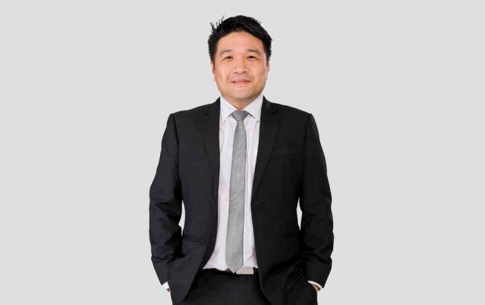 A portrait medium-shot of Sunway Group chief information officer Kevin Khoo Min Sinn, smirking at the camera with both hands tucked into his pants pocket, dressed in clasic black suit and a grey tie