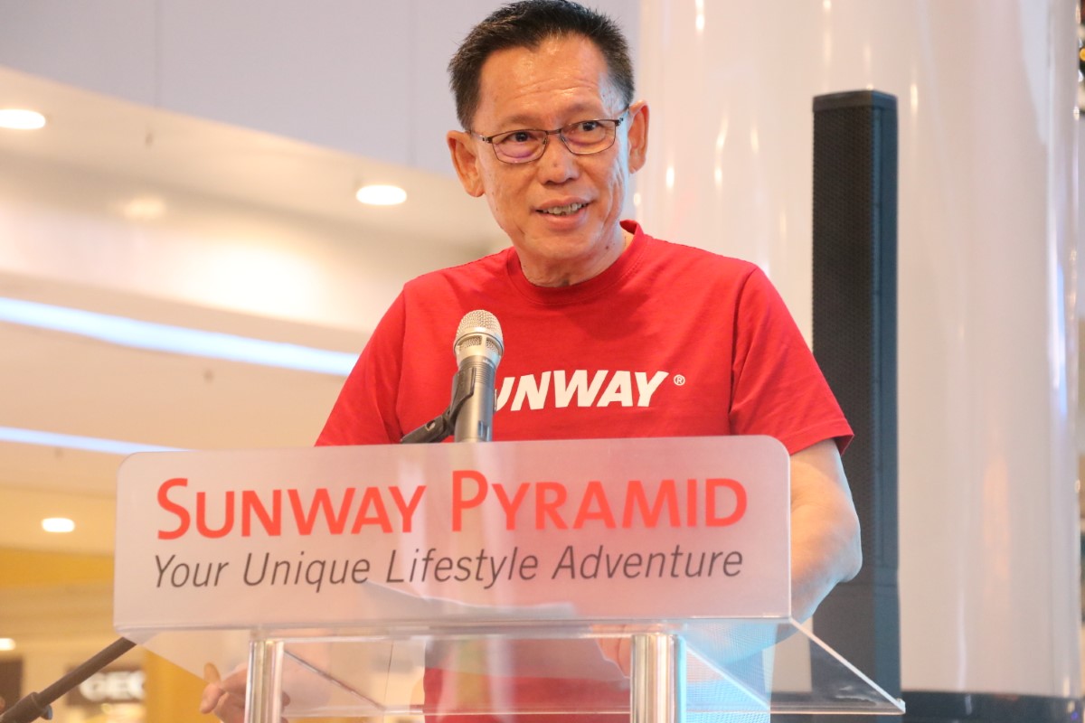A mid-wide shot of Sunway Malls & Theme Parks chief executive officer HC Chan in a signature red Sunway tee and horn-rimmed glasses, speaking indoors behind a Sunway Pyramid podium
