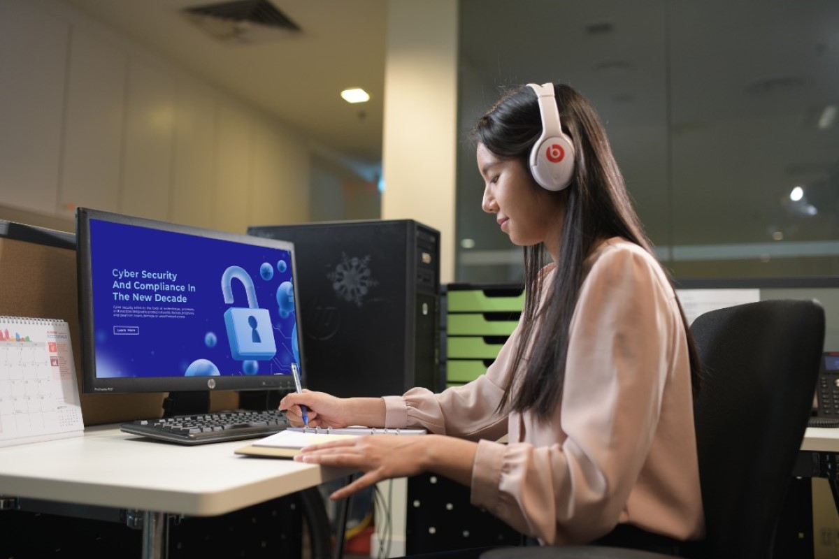 A medium shot of a side profile of a female Sunwayian with long hair in white headphones attending a virtual training, jotting down notes with a pen and notebook.