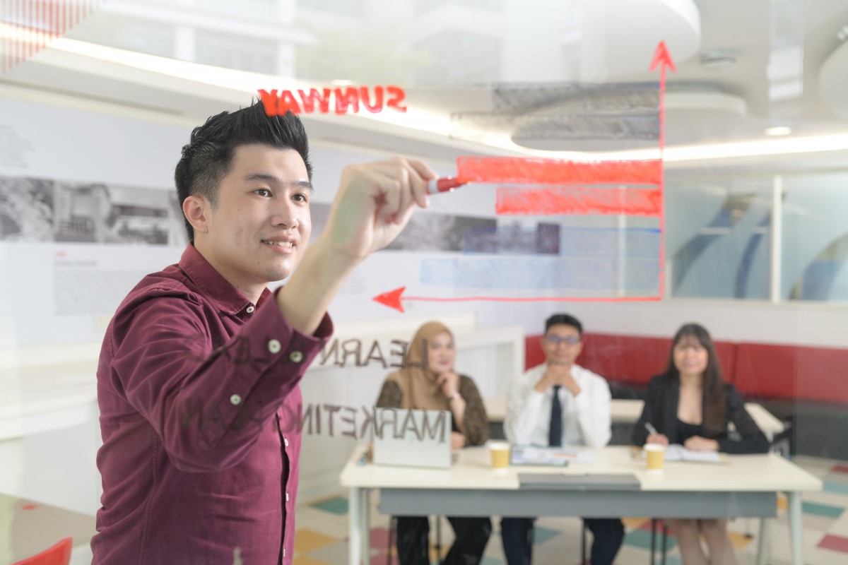 A medium shot of a male Sunwayian in a maroon button-up drawing a bar graph on a transparent board, as three other Sunwayians remain focused on him from a distance away.