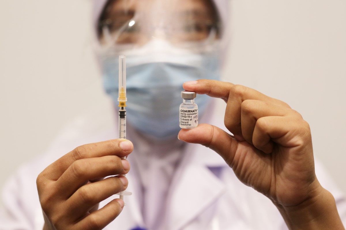 A close-up of a frontline worker or lab physician in surgical mask and full-face shield holding up a covered syringe in one hand and a bottle of COMIRNATY vaccine solution in the other with bare hands.