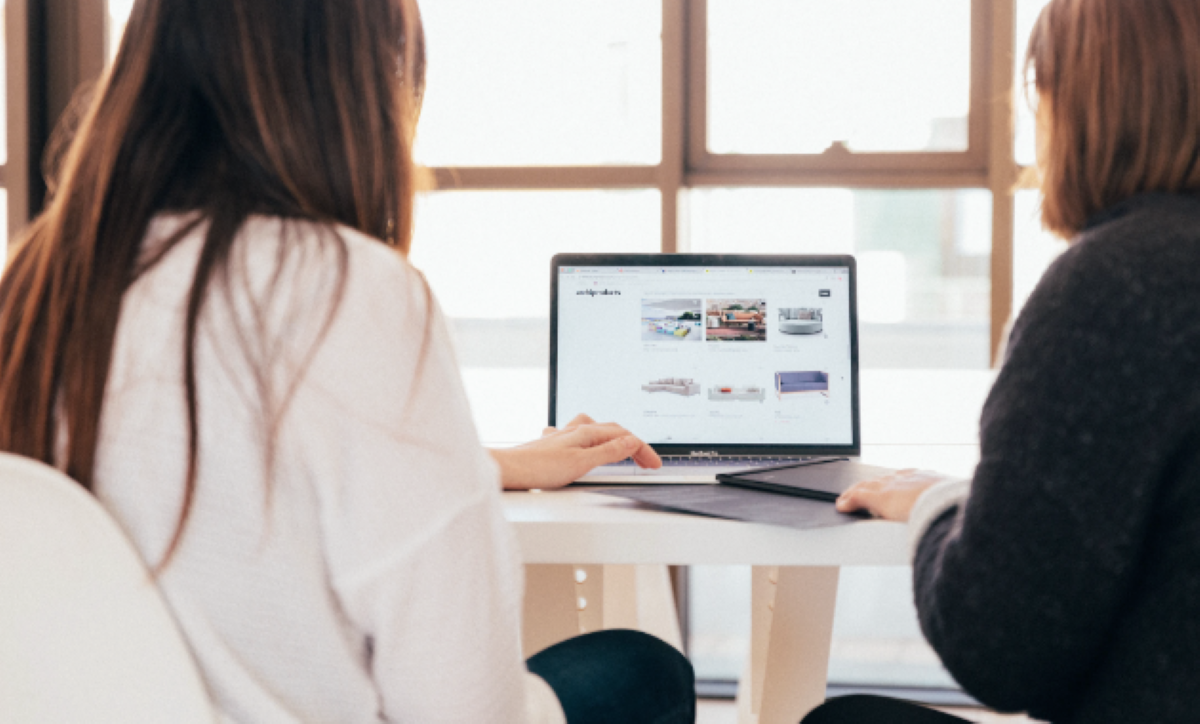 A mid wide shot of two female brunettes sitting side by side, facing an open laptop on a table, in front of window panes, surrounded by bright daylight.