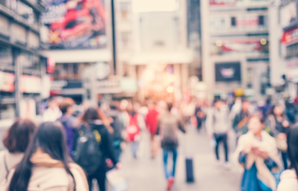 A blurred wide shot of a bustling street in a city at daytime, with people’s faint silhouettes and surrounding buildings outlining the frame.