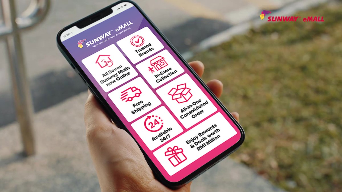 A close-up of a left hand holding up a smartphone displaying Sunway eMall homepage.