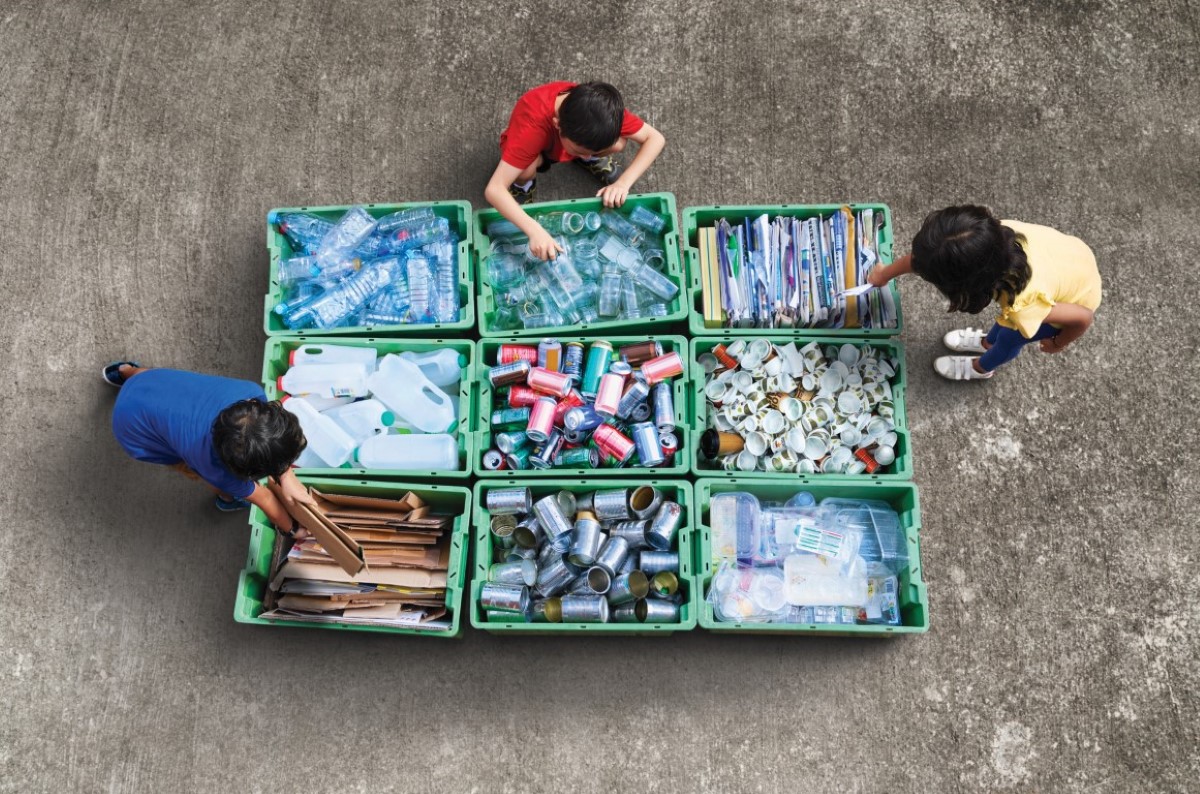 A wide high angle shot of nine recycle bins separating waste materials in an organised manner, with three persons dressed in blue, red and yellow tops respectively, rummaging through the bins by the side.