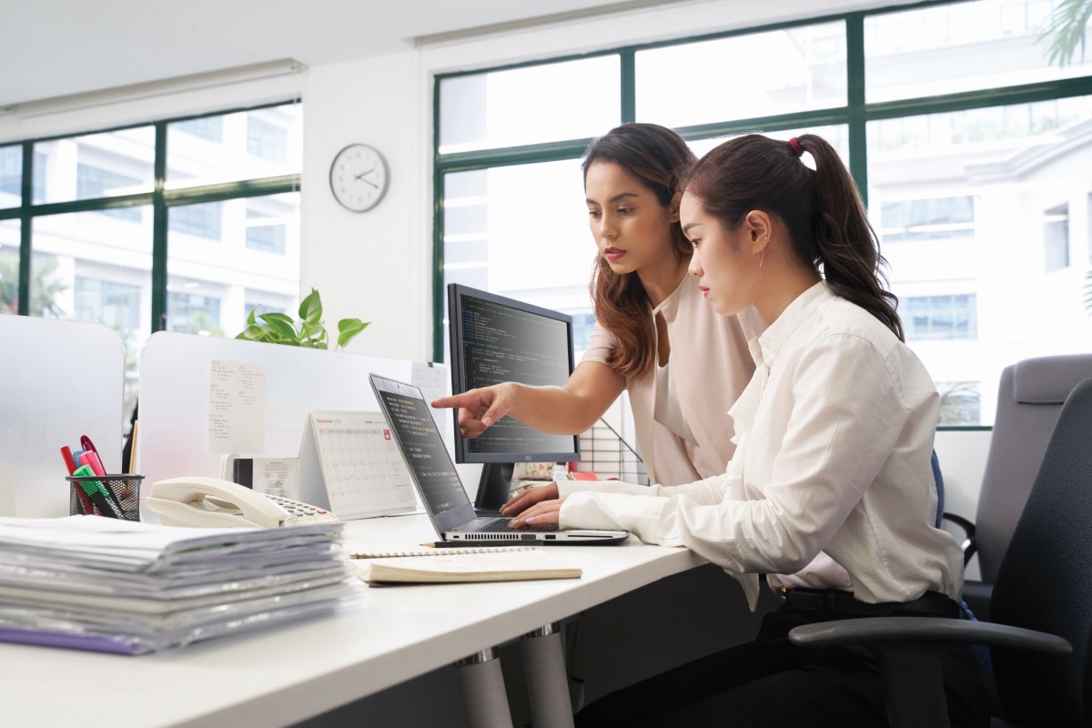 A medium full-shot featuring the side profiles of two office ladies with their eyes fixated on an open laptop on the desk in front of them, surrounded by a clean and white office space with bright daylight pouring in from the nearby windows.