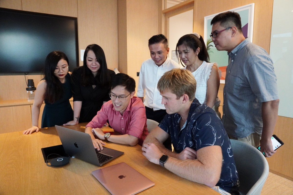 A medium full shot of Trabble’s team of many in a warm-toned conference room – all standing behind founders Low Jian Liang and Rys Bilinski, all eyes fixated on an open laptop facing them.