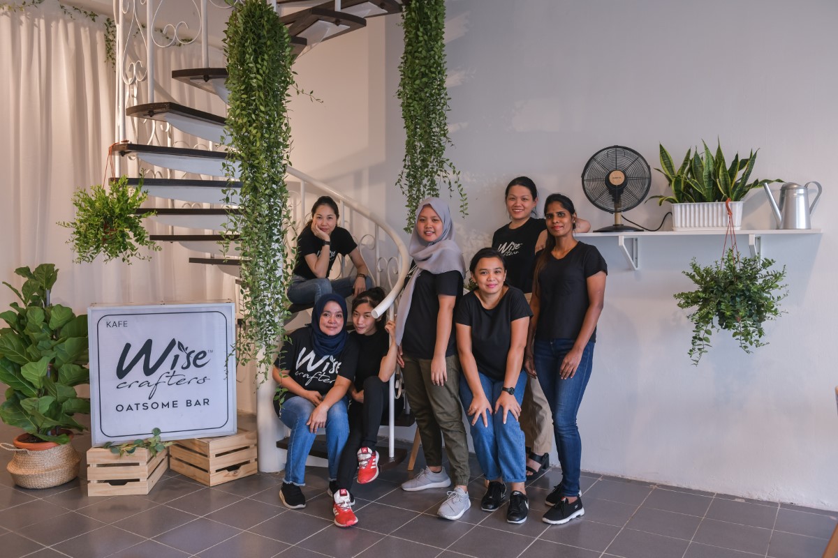 A full shot of Wise Crafters’ large team comprising all females – all wearing their signature black tee shirt, posing around a white spiral staircase amidst an open, clean white space, led by founders Genie Hor and Samantha Tan.