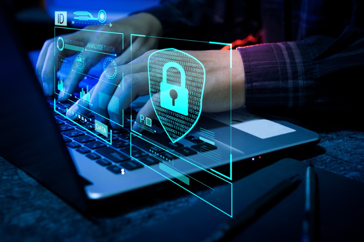 A close-up of image of pair of male hands typing away on a laptop’s keyboard, with the laptop screen’s blue light illuminating the visual, some cyan hologram with a padlock and nondescript codes floating above the keyboard