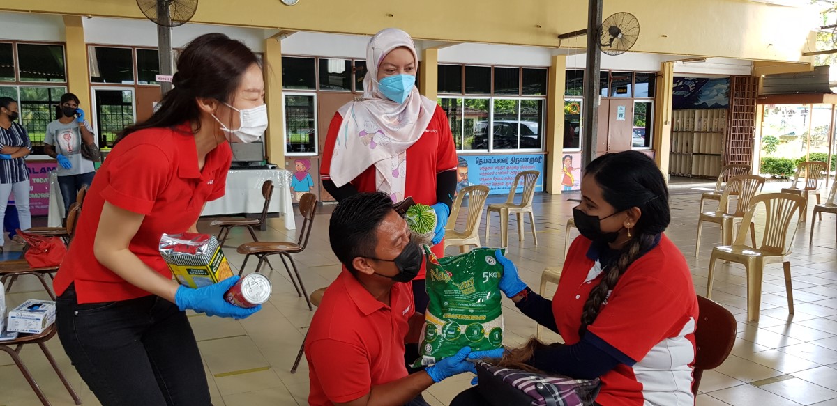 A medium landscape shot of three Sunwayians in our signature red polo tee and big smiles beneath their respective face masks presenting essential groceries to an Indian lady at a spacious and deserted hall