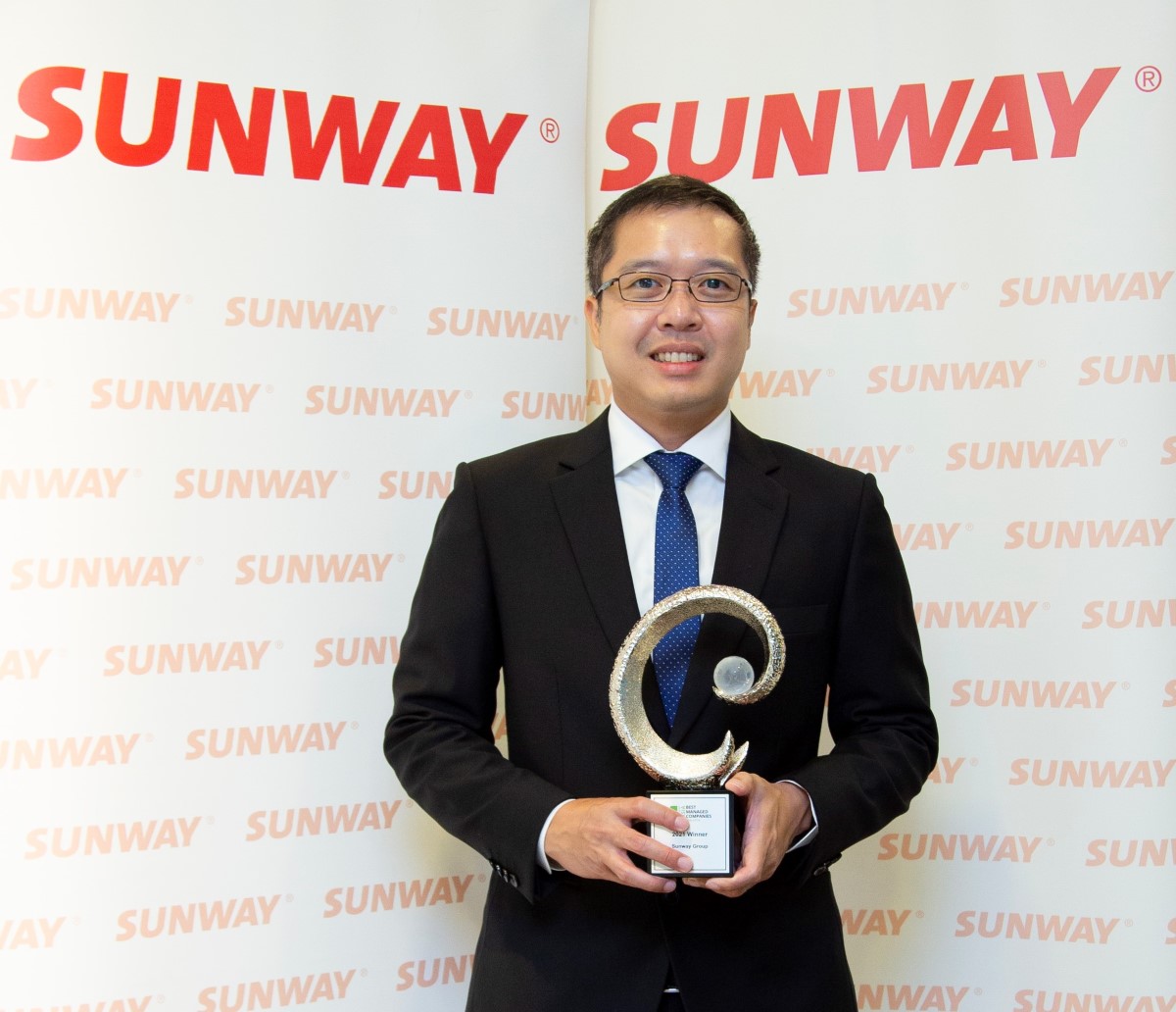 : A medium shot of Evan Cheah who is Executive Vice President of President’s Office at Sunway Berhad, dressed impeccably smart in black suit, white button up and blue tie, holding Malaysia’s Best Managed Companies 2021 trophy conferred by global consulting firm Deloitte, fronting a simple red-and-white Sunway backdrop