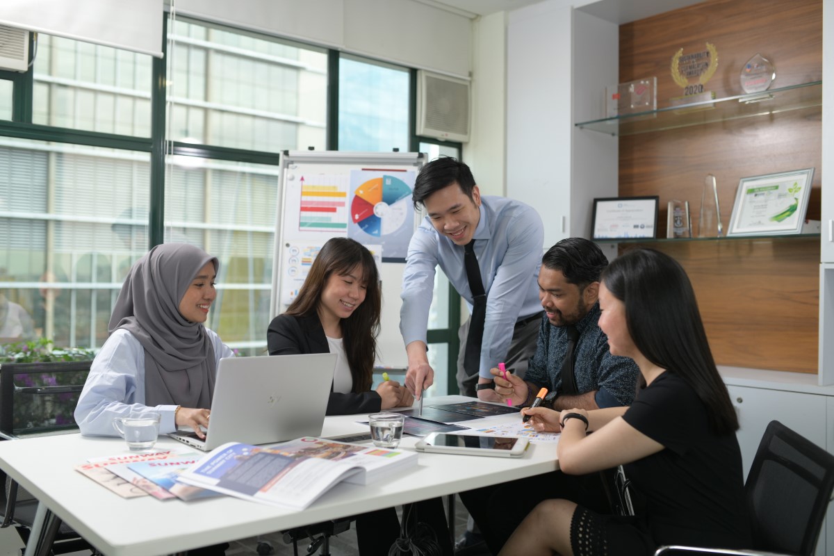 A medium landscape shot of five Sunwayians engaged in an interactive engagement, four seated while one remain standing to facilitate the discussion, the table filled with necessary materials and tools while an informative whiteboard can be seen in the background