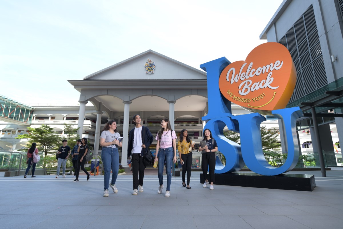 Sunway students of different races carrying bags and books walking to class, with the “I Love SU” sign and Sunway College building in the background.