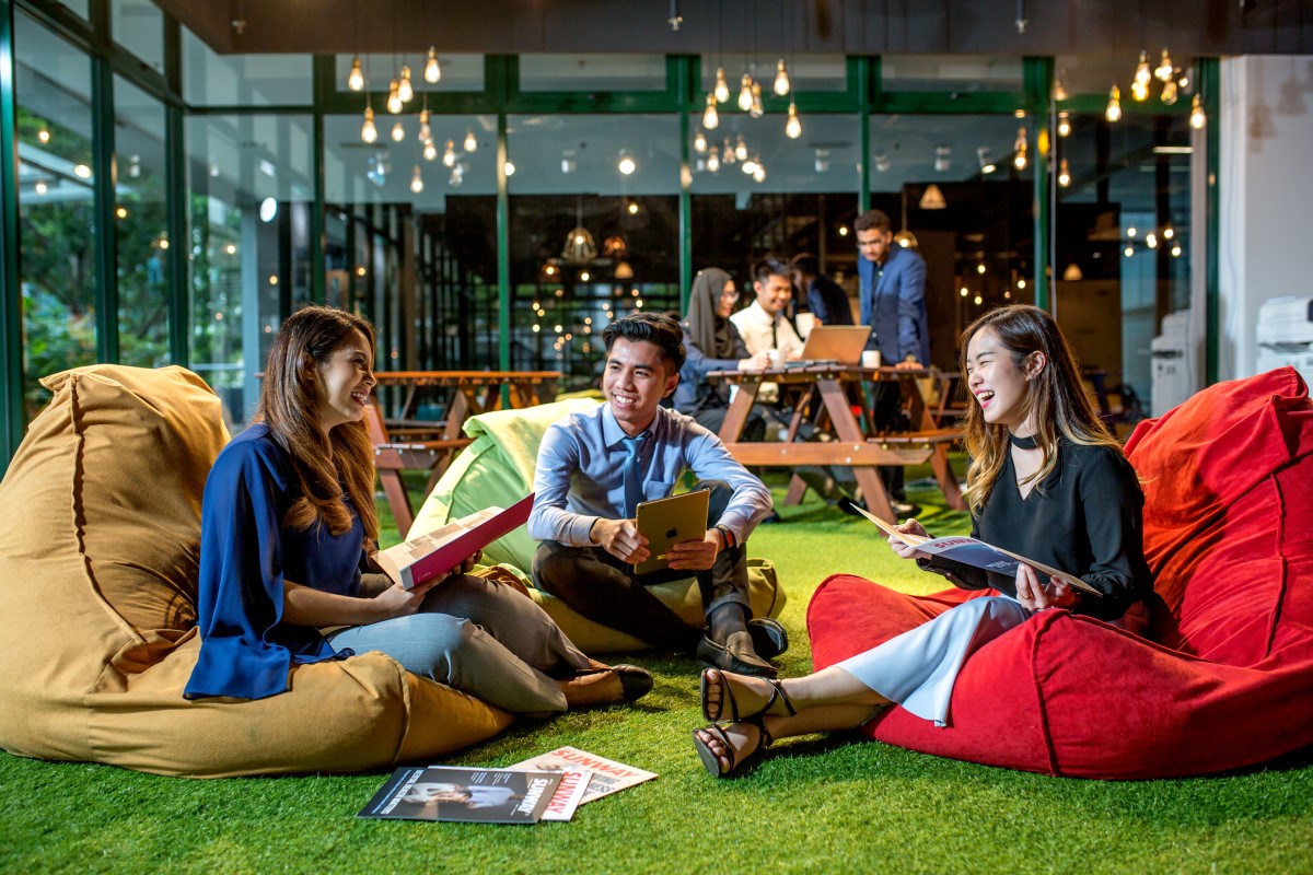 A full landscape shot of three Sunwayians interacting jovially while sitting on bean bags on the grassy ground while another three Sunwayians remain fixated on an open laptop on a picnic table in the background