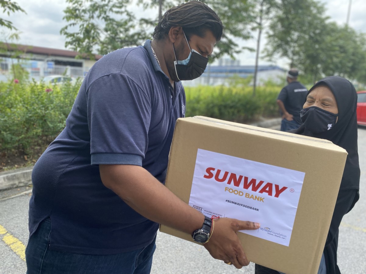 A medium full landscape shot of a Human Resources for Rural Areas (DHRRA) representative in face mask handing over a big boxful of essential groceries labelled “Sunway Food Bank” to a Malay lady in face mask during Sunway Group’s #SunwayforGood Food Bank National Day campaign in 2021