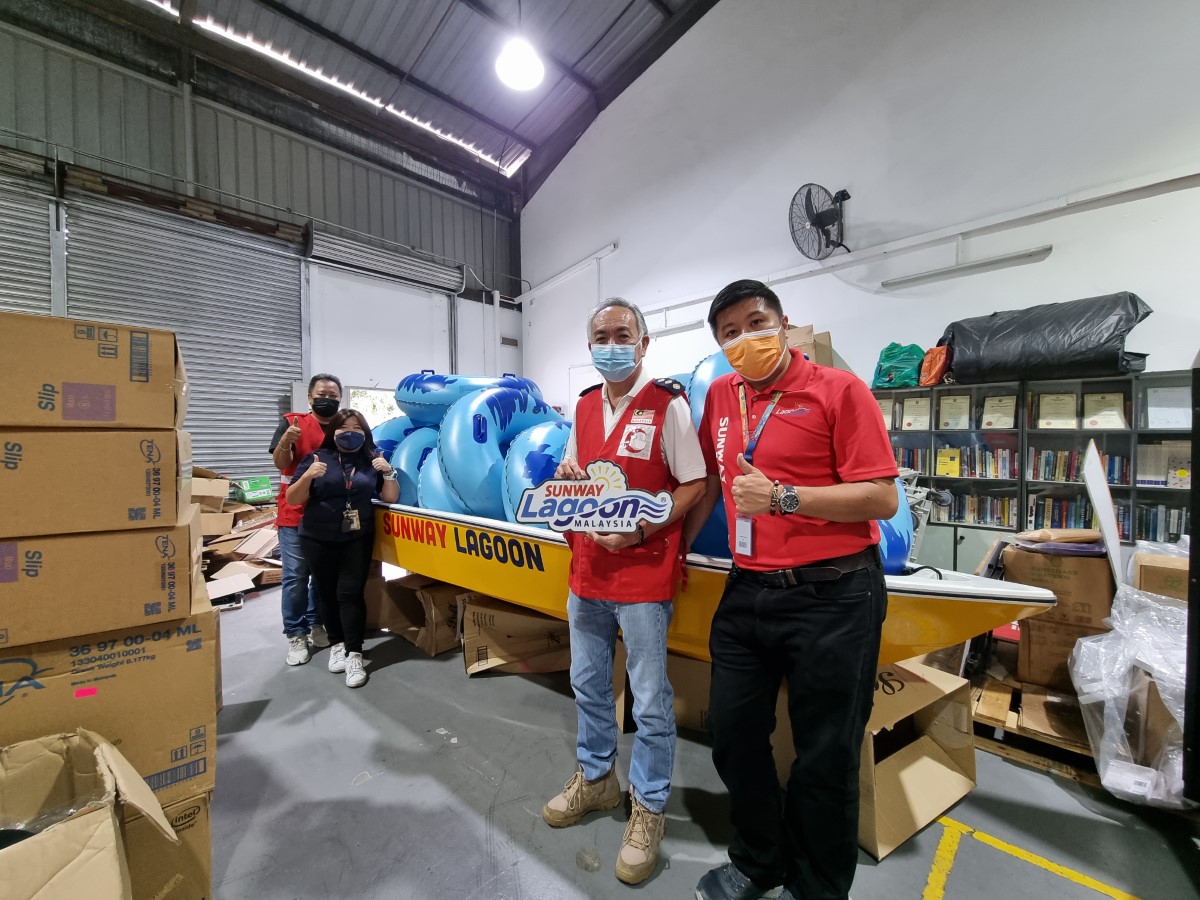 A full landscape shot of Sunwayians in Sunwayians in their signature red polo tee and face masks alongside representative members of the Malaysian Red Crescent Society (MRC) in unifron and face mask posing amidst a warehouse full of necessities to aid flood victims in affected Selangor areas