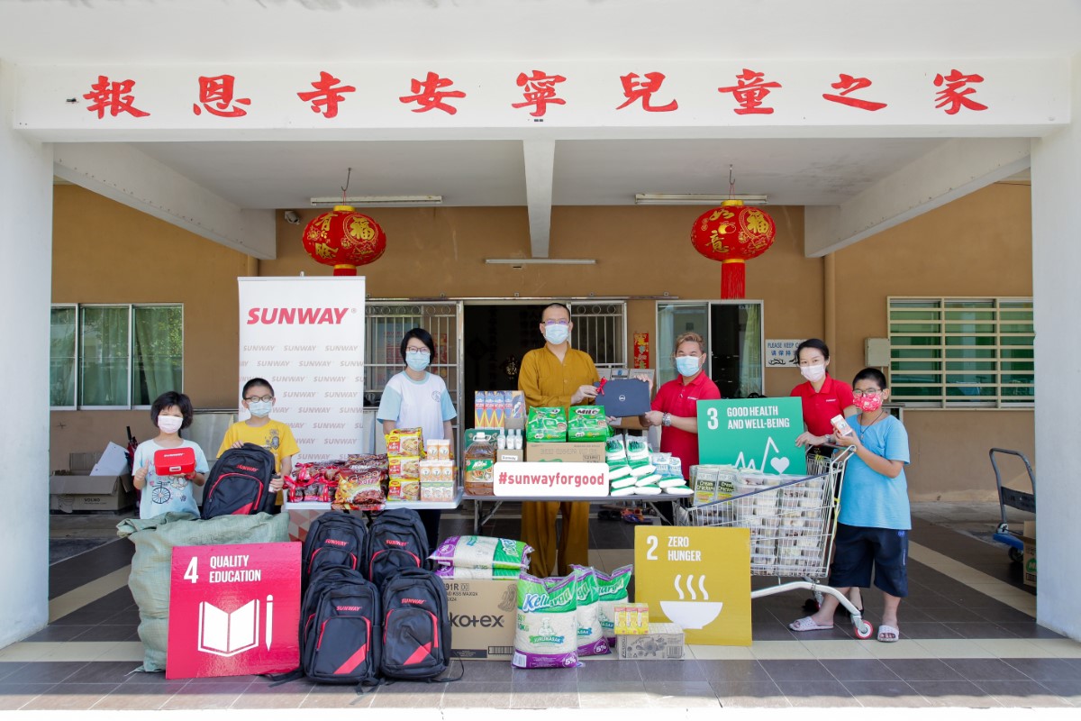 A full landscape shot of Sunwayians in their signature red polo tee and face masks posing alongside beneficiaries of multiracial children home in face masks behind a mountain of essential groceries and school necessities, as part of Sunway Group’s Chinese New Year cheer programme