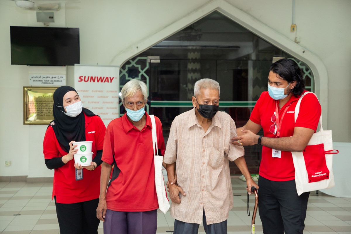 A medium full landscape shot of Sunwayians in their signature red polo tee and face masks assisting the elderly in face masks in front of a mosque during Sunway Group’s Raya cheer initiative in 2021