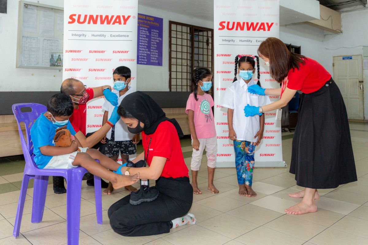 A full landscape shot of three Sunwayians in signature red polo top and face masks, each attending to a child fitted to their new school uniforms in an open hall