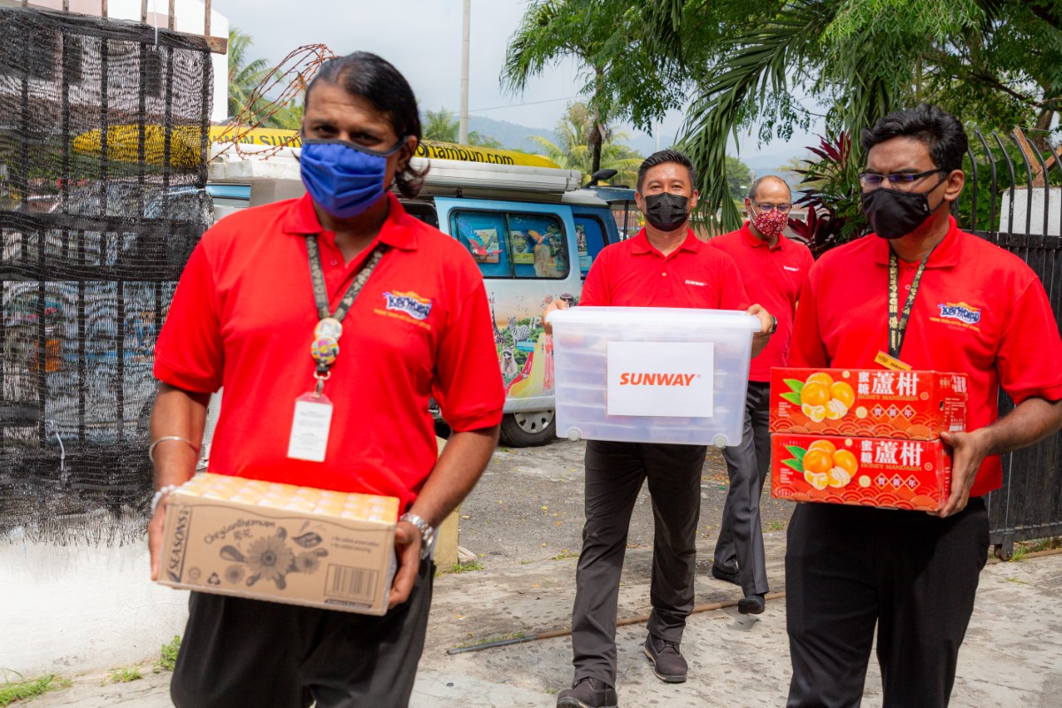 A medium full landscape shot of four Sunwayians - each in signature red polo tee and face masks carrying boxes after boxes of necessities and festive essentials from a Sunway Lost World of Tambun van in the background
