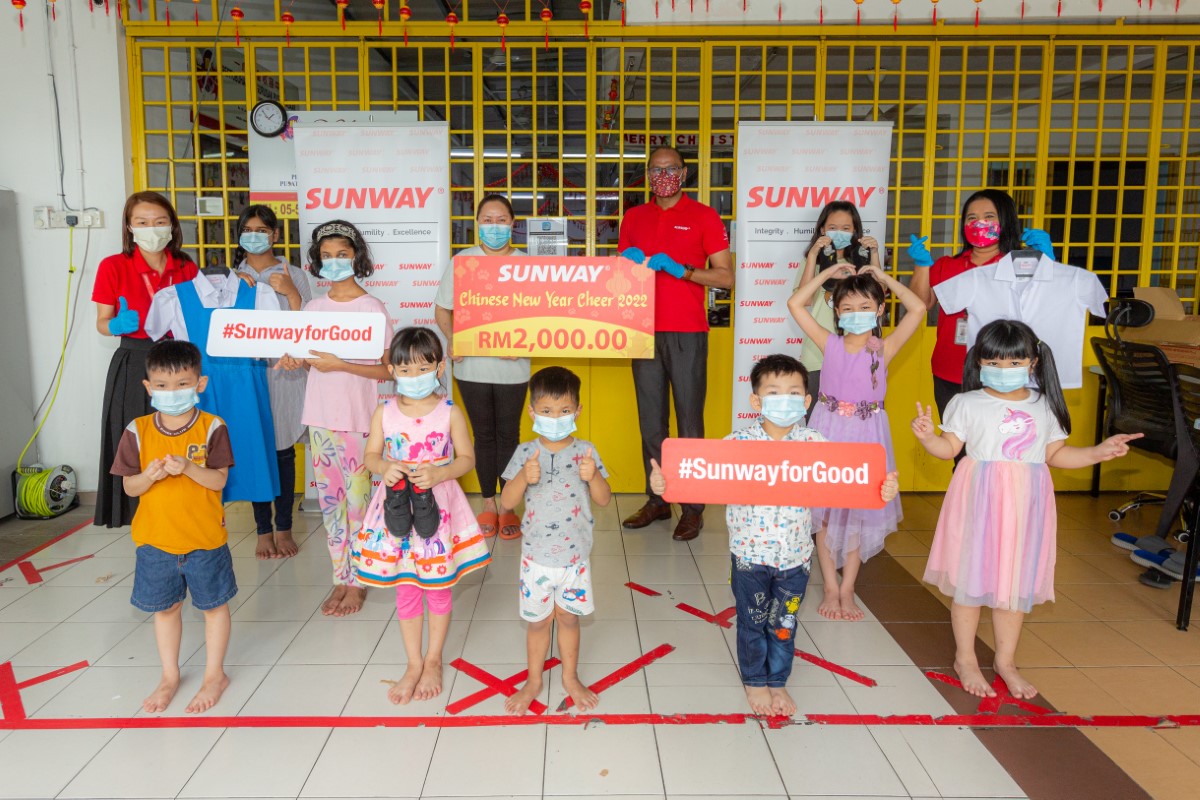 A full landscape shot of Sunwayians in signature red polo tee amidst children presenting the home representative with a RM2,000 mock cheque alongside school essentials as part of Sunway Group’s Chinese New Year Cheer in 2022