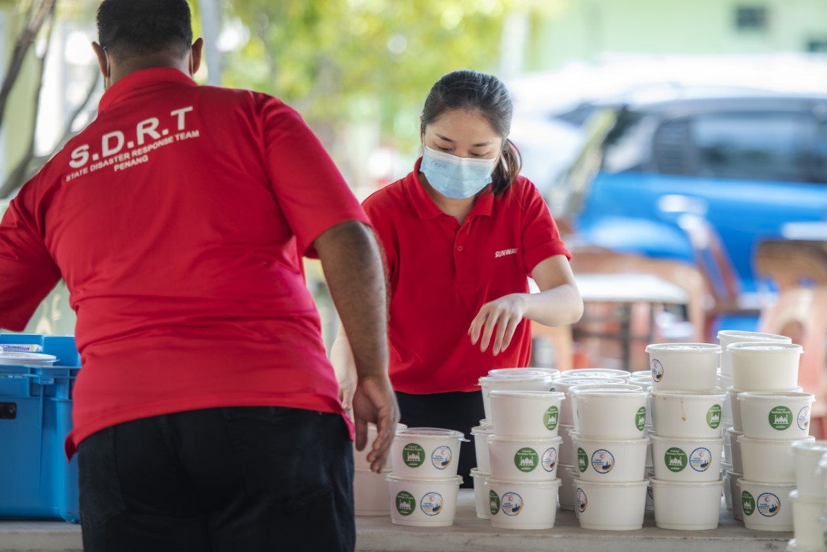 A medium landscape shot of a female Sunwayian in signature red polo tee and face mask stacking containers of bubur lambuk alongside a member of our partner organisation during Raya Cheer 2021